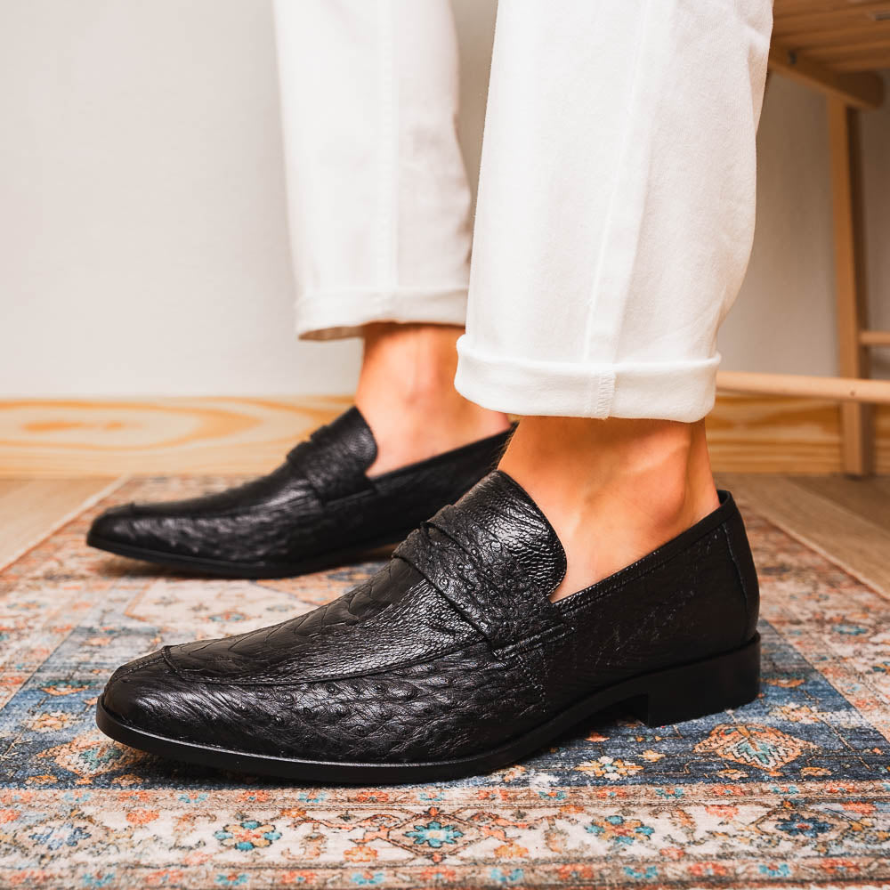 The Fangio Black by Marco Di Milano: a pair of black textured leather loafers, one upright and one sole-up, displaying the brand on the tan sole—luxury men's footwear to elevate any look.