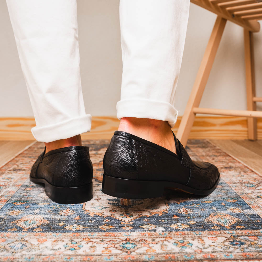 The Fangio Black by Marco Di Milano: a pair of black textured leather loafers, one upright and one sole-up, displaying the brand on the tan sole—luxury men's footwear to elevate any look.