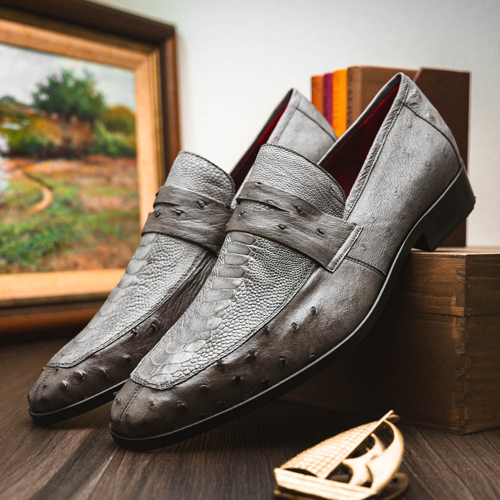 The Fangio Serpentine Gray by Marco Di Milano features gray textured leather in classic penny loafer style, with detailed stitching and tan leather soles—one shoe upright, one on its side—to showcase refined craftsmanship.