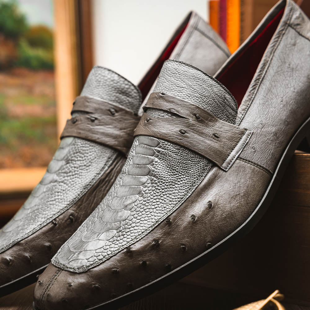 The Fangio Serpentine Gray by Marco Di Milano features gray textured leather in classic penny loafer style, with detailed stitching and tan leather soles—one shoe upright, one on its side—to showcase refined craftsmanship.
