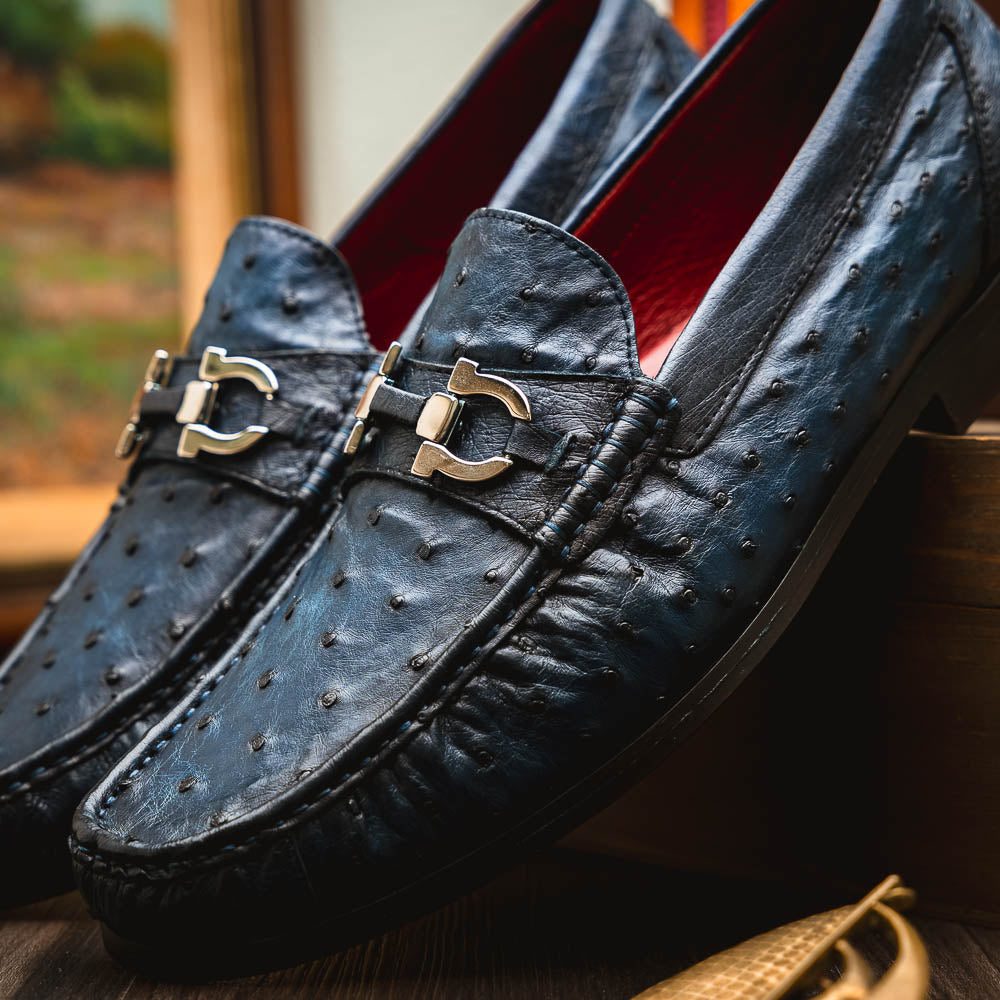 A pair of luxury men's shoes: Ferrioni Navy loafers by Marco Di Milano in genuine ostrich leather, featuring classic bit detail. One shoe is upright, the other shows the sole with "Marco Di Milano" branding.