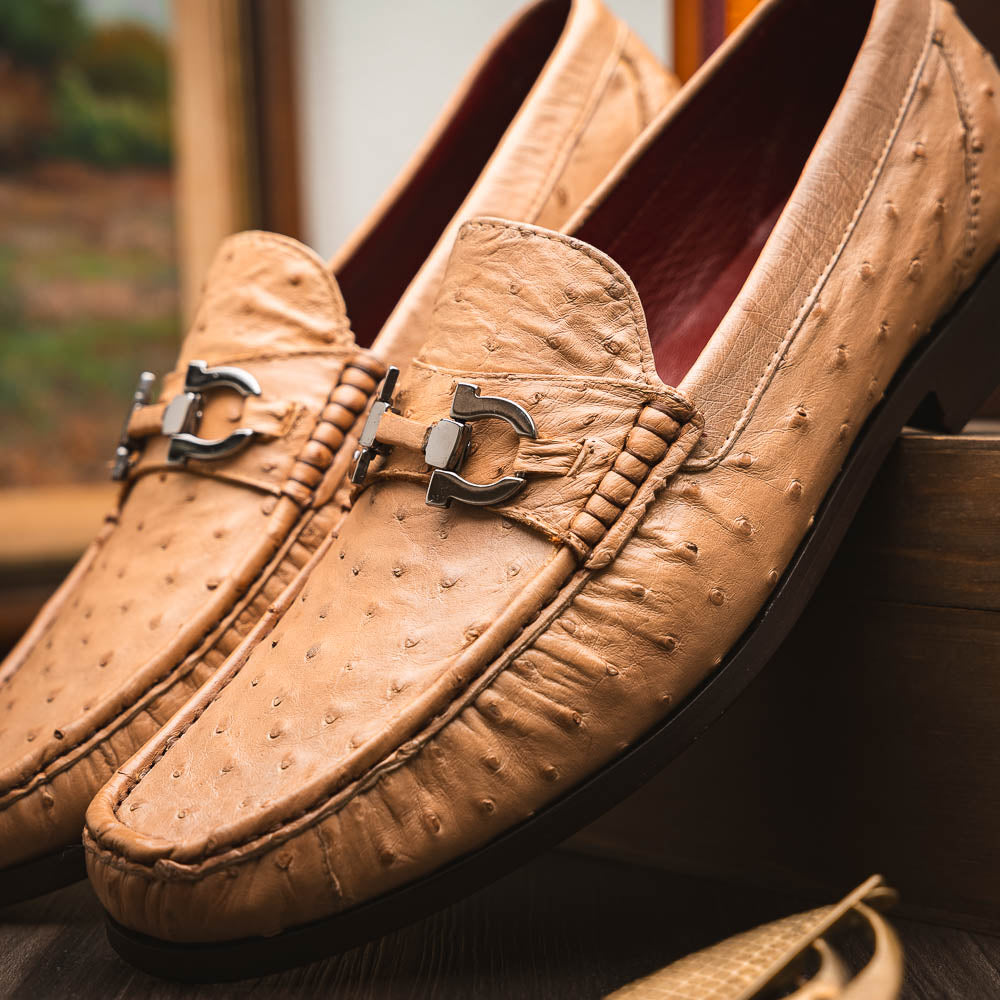 The Marco Di Milano Ferrioni Orix is a pair of genuine beige ostrich loafers with a classic bit buckle, one upright and one on its side showing the insole logo—timeless luxury men's footwear.