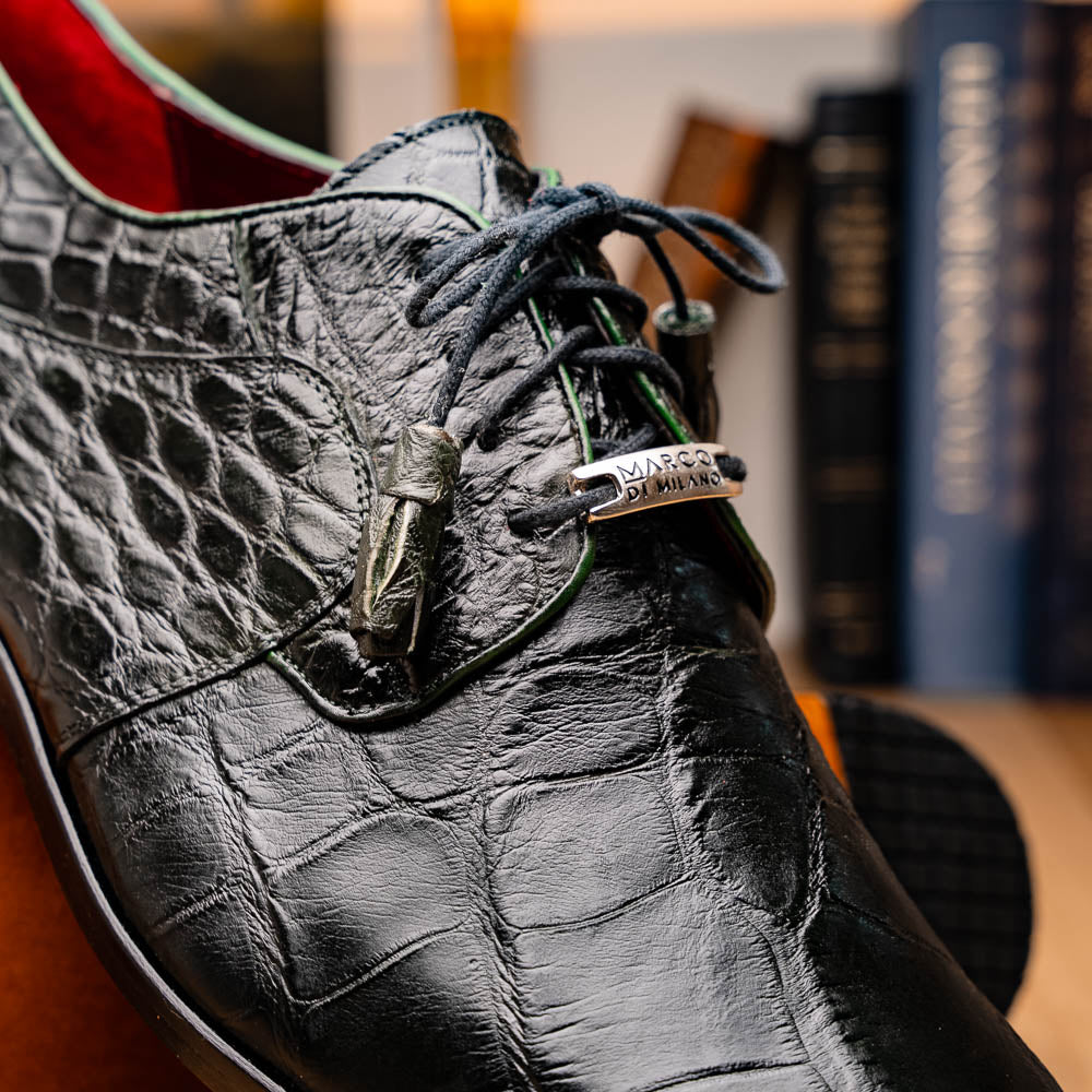 A pair of Fitipaldi Olive luxury footwear by Marco Di Milano in black crocodile leather—one shoe upright, the other showing a tan sole with "Marco Di Milano" embossed—crafted with hand-painted finishes for an elegant touch.