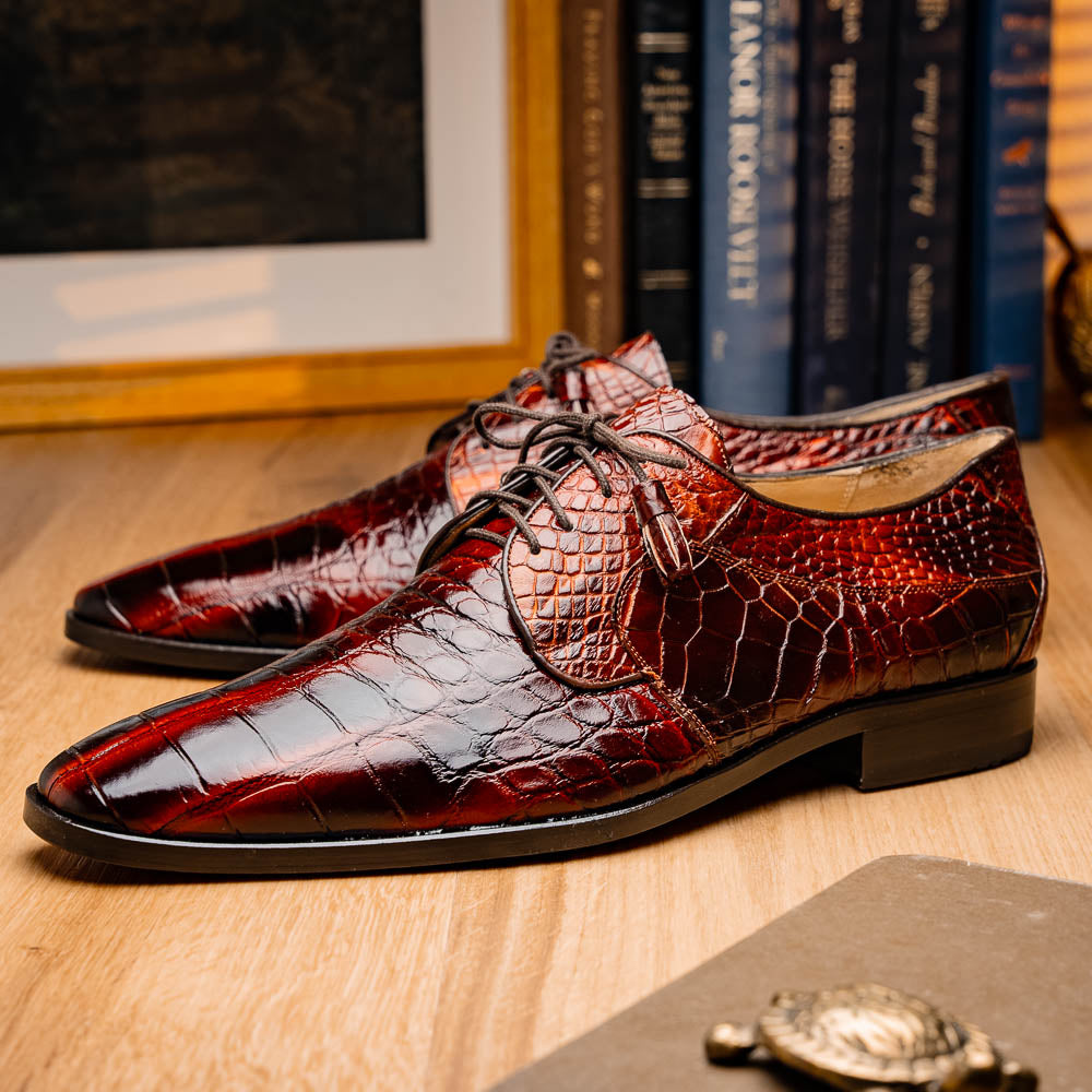 A pair of Fitipaldi Rustic Cognac shoes by Marco Di Milano in brown crocodile-patterned leather—one upright, one displaying the sole with branding—showcases exquisite hand-painted finishes, epitomizing luxury footwear.