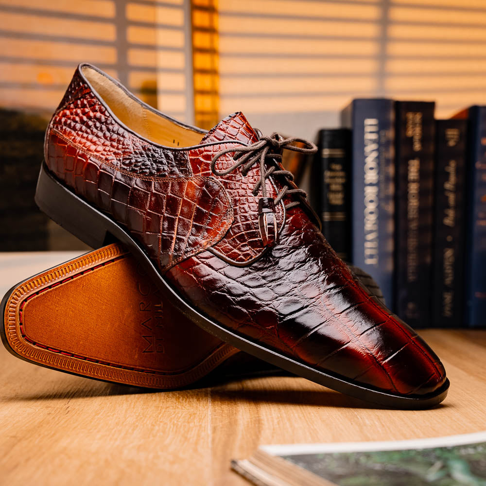 A pair of Fitipaldi Rustic Cognac shoes by Marco Di Milano in brown crocodile-patterned leather—one upright, one displaying the sole with branding—showcases exquisite hand-painted finishes, epitomizing luxury footwear.