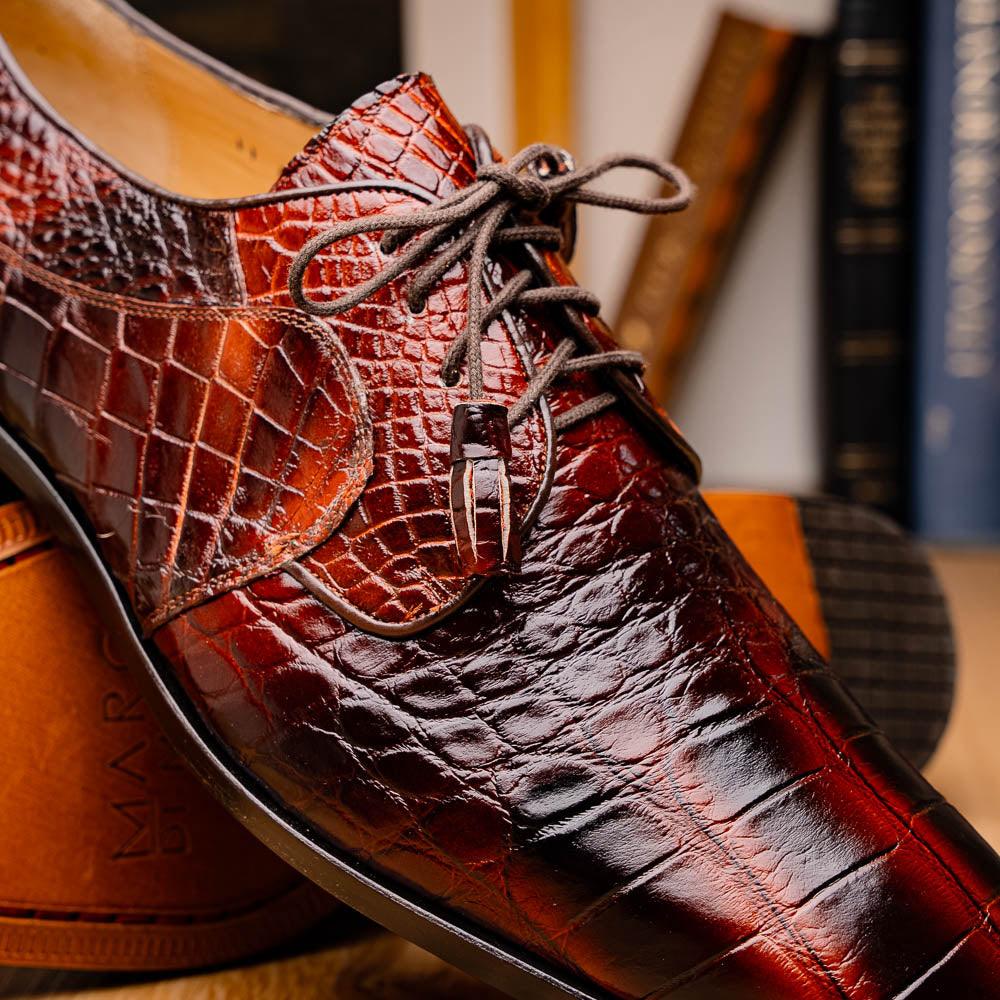 A pair of Fitipaldi Rustic Cognac shoes by Marco Di Milano in brown crocodile-patterned leather—one upright, one displaying the sole with branding—showcases exquisite hand-painted finishes, epitomizing luxury footwear.