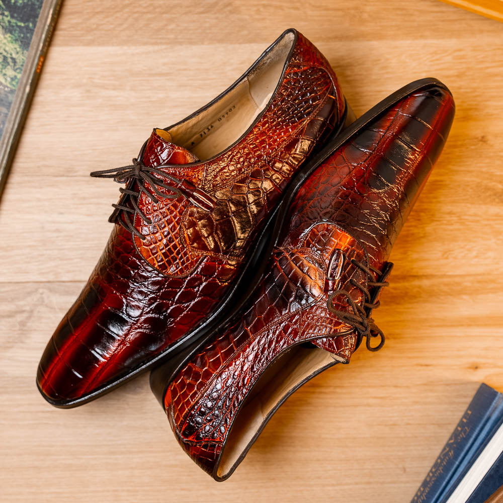 A pair of Fitipaldi Rustic Cognac shoes by Marco Di Milano in brown crocodile-patterned leather—one upright, one displaying the sole with branding—showcases exquisite hand-painted finishes, epitomizing luxury footwear.