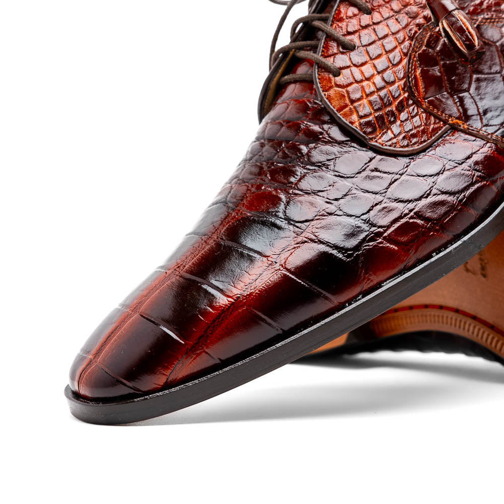 A pair of Fitipaldi Rustic Cognac shoes by Marco Di Milano in brown crocodile-patterned leather—one upright, one displaying the sole with branding—showcases exquisite hand-painted finishes, epitomizing luxury footwear.