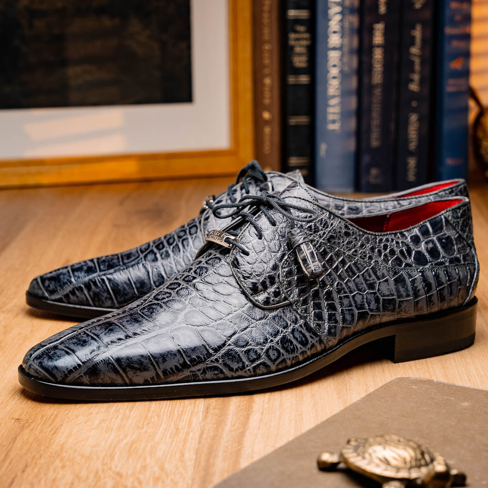A pair of gray crocodile-pattern luxury dress shoes, the Fitipaldi Rustic Gray by Marco Di Milano, are shown—one upright and one on its side—to display a tan sole marked "Marco Di Milano." Hand-painted finishes showcase exquisite craftsmanship.