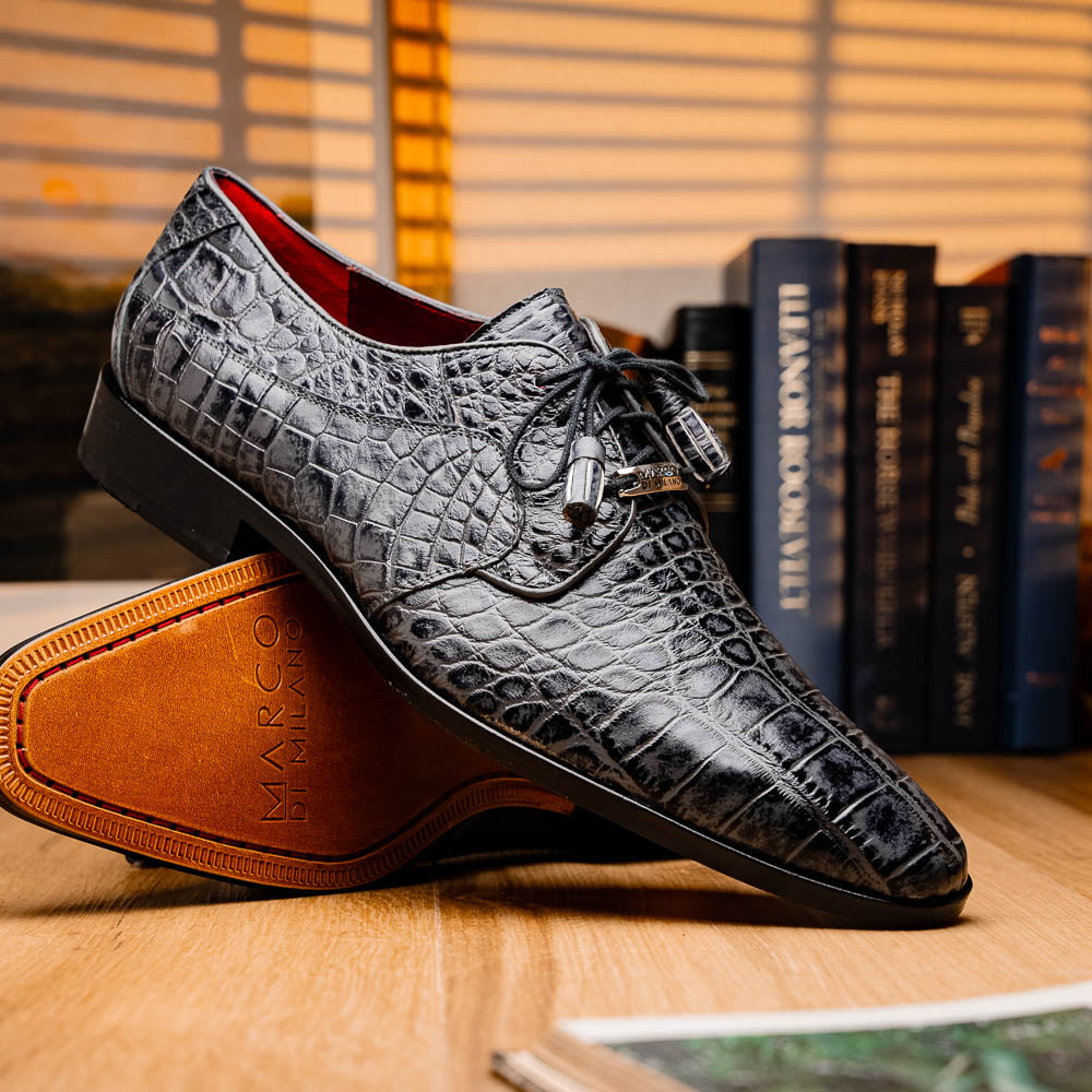 A pair of gray crocodile-pattern luxury dress shoes, the Fitipaldi Rustic Gray by Marco Di Milano, are shown—one upright and one on its side—to display a tan sole marked "Marco Di Milano." Hand-painted finishes showcase exquisite craftsmanship.