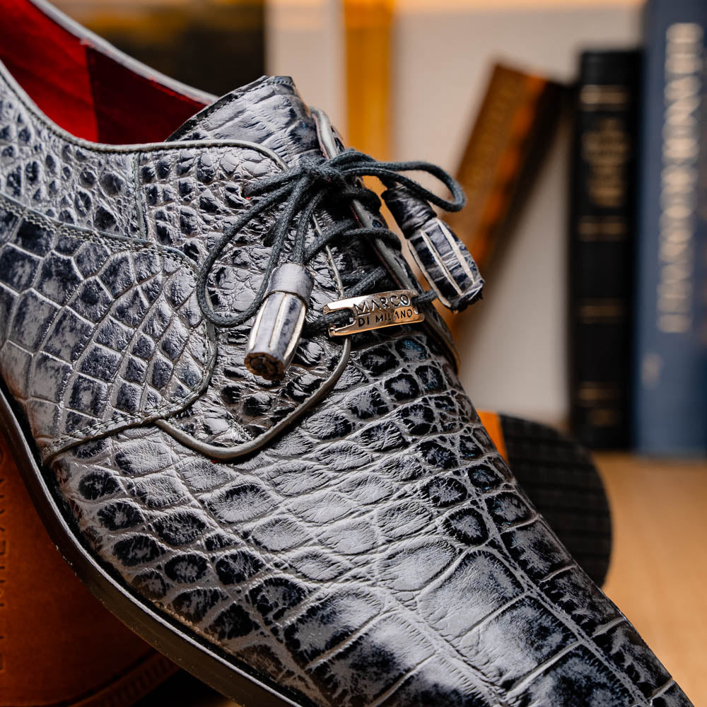 A pair of gray crocodile-pattern luxury dress shoes, the Fitipaldi Rustic Gray by Marco Di Milano, are shown—one upright and one on its side—to display a tan sole marked "Marco Di Milano." Hand-painted finishes showcase exquisite craftsmanship.