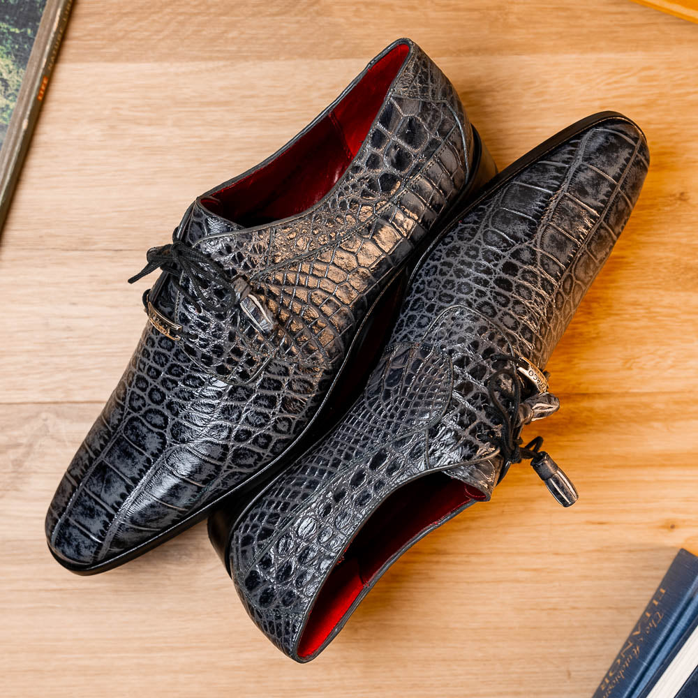 A pair of gray crocodile-pattern luxury dress shoes, the Fitipaldi Rustic Gray by Marco Di Milano, are shown—one upright and one on its side—to display a tan sole marked "Marco Di Milano." Hand-painted finishes showcase exquisite craftsmanship.