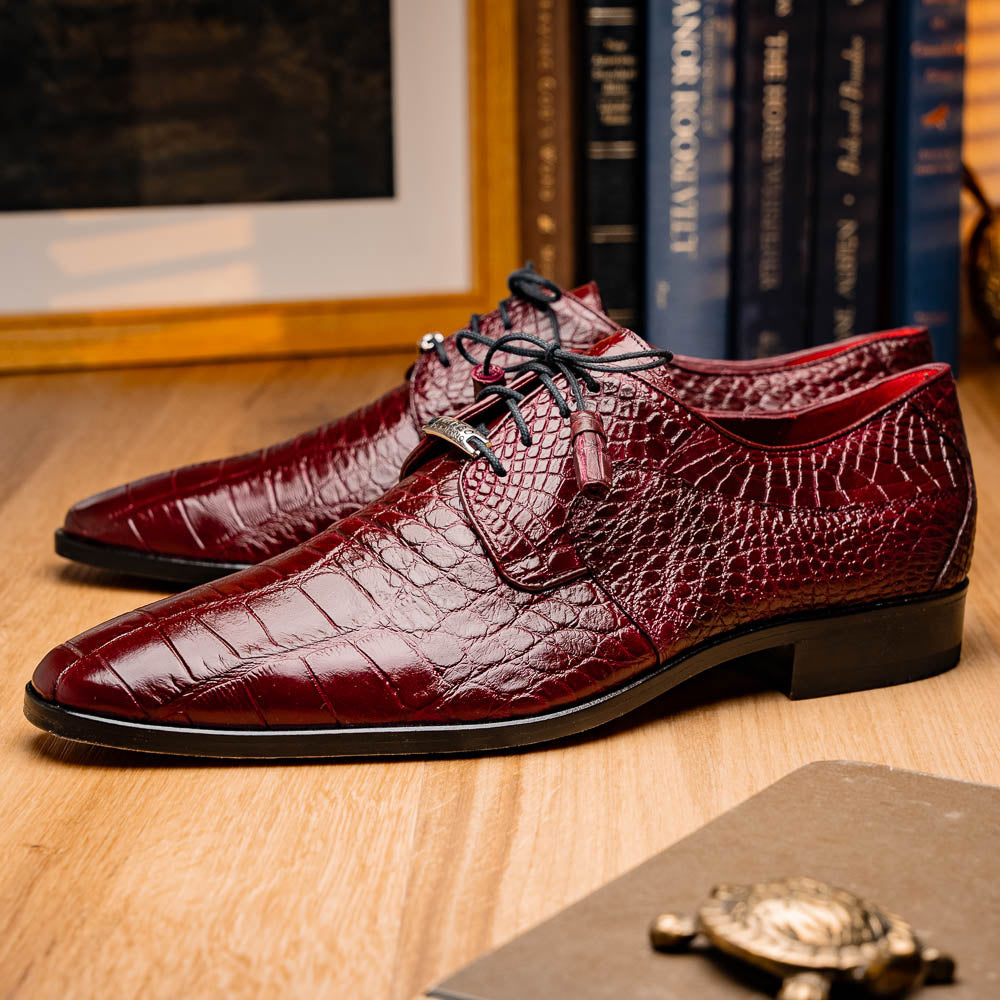 The Fitipaldi Wine by Marco Di Milano is a burgundy crocodile-pattern leather dress shoe with black laces, shown on its side to reveal the tan sole and logo—highlighting hand-painted details and luxury craftsmanship.