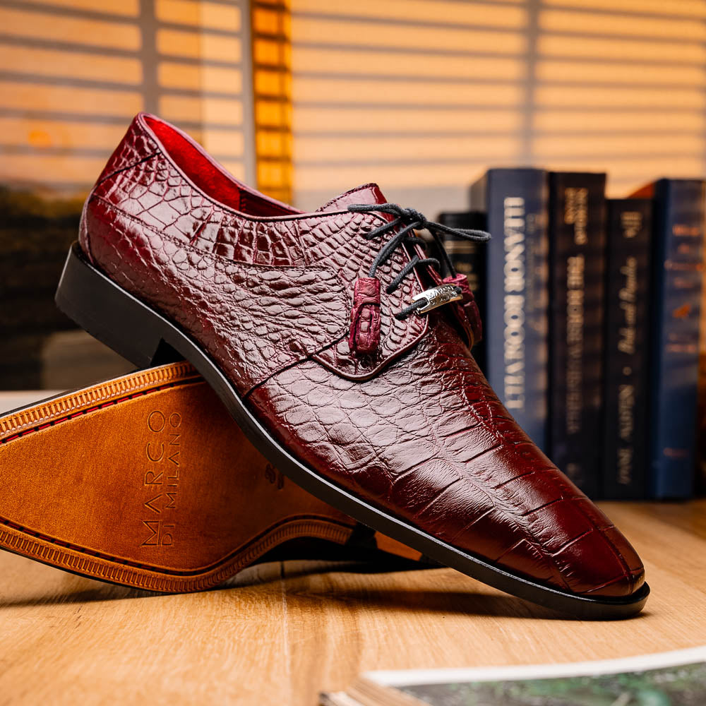 The Fitipaldi Wine by Marco Di Milano is a burgundy crocodile-pattern leather dress shoe with black laces, shown on its side to reveal the tan sole and logo—highlighting hand-painted details and luxury craftsmanship.