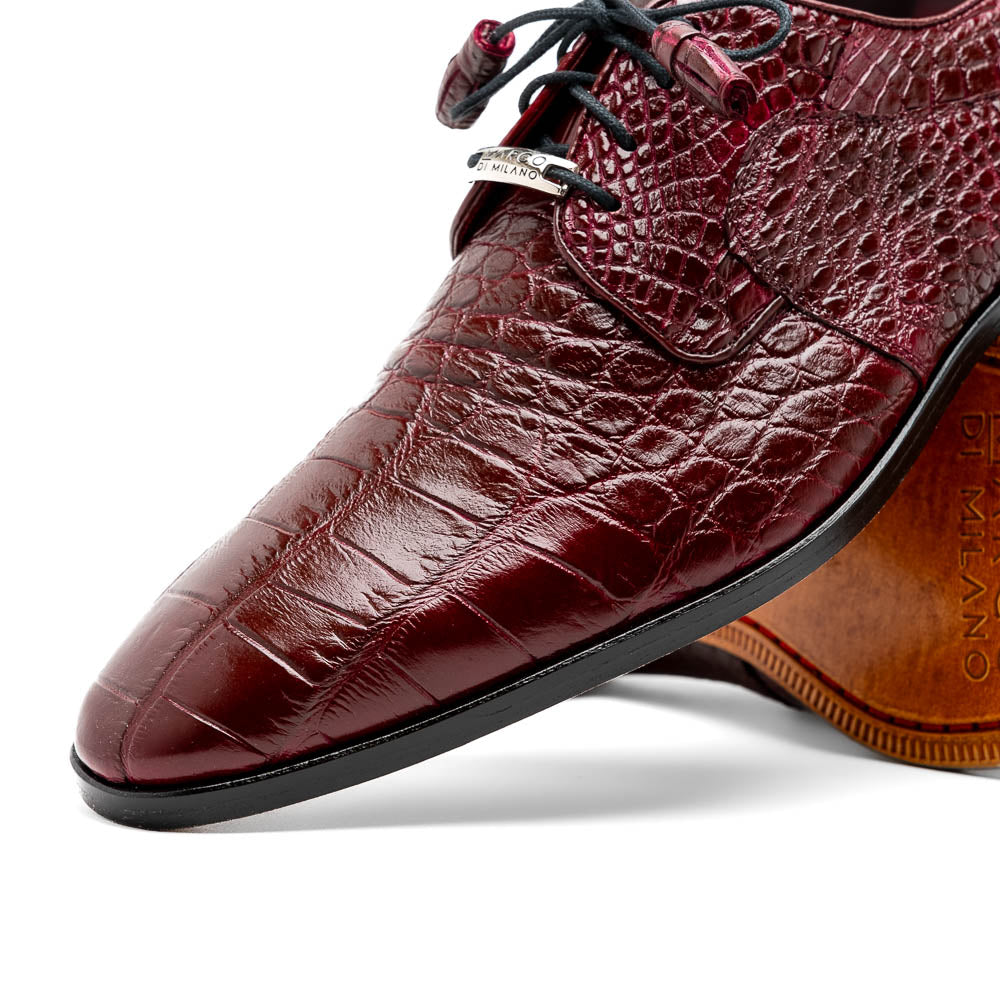 The Fitipaldi Wine by Marco Di Milano is a burgundy crocodile-pattern leather dress shoe with black laces, shown on its side to reveal the tan sole and logo—highlighting hand-painted details and luxury craftsmanship.