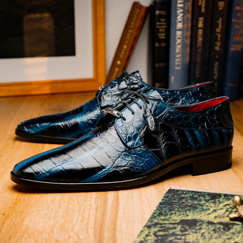 Fitipaldi Rustic Navy by Marco Di Milano: a pair of blue leather men’s dress shoes with a crocodile pattern, black soles, and lace-up fronts; one luxury shoe is shown on its side to display the tan sole.