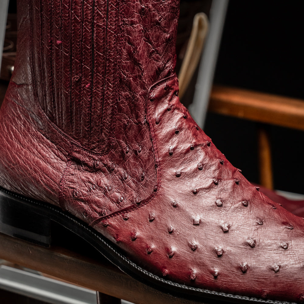 Introducing Giorgio Wine by Marco Di Milano: a pair of luxurious men's boots handcrafted in genuine burgundy ostrich leather, featuring a distinctive textured pattern, side zipper, elastic panel, and a sleek black sole—sophisticatedly presented against a white background.