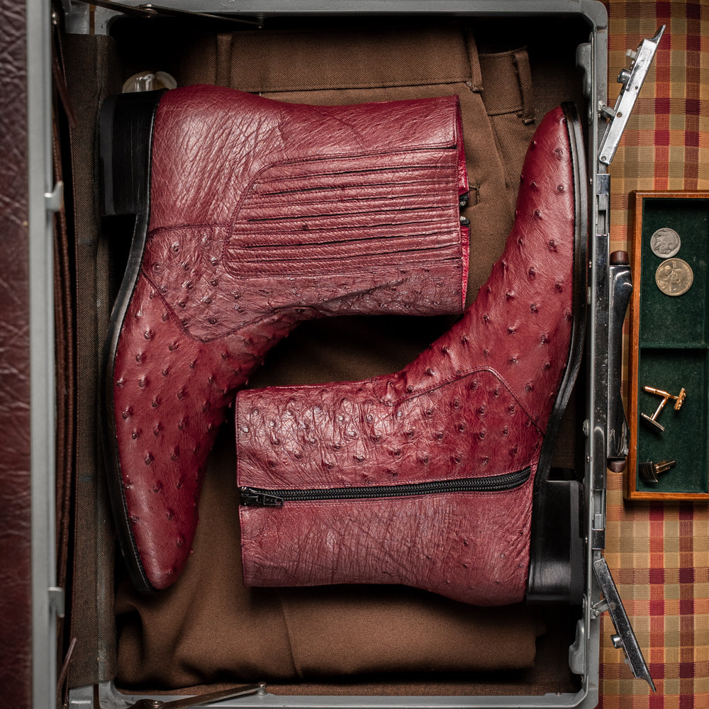Introducing Giorgio Wine by Marco Di Milano: a pair of luxurious men's boots handcrafted in genuine burgundy ostrich leather, featuring a distinctive textured pattern, side zipper, elastic panel, and a sleek black sole—sophisticatedly presented against a white background.