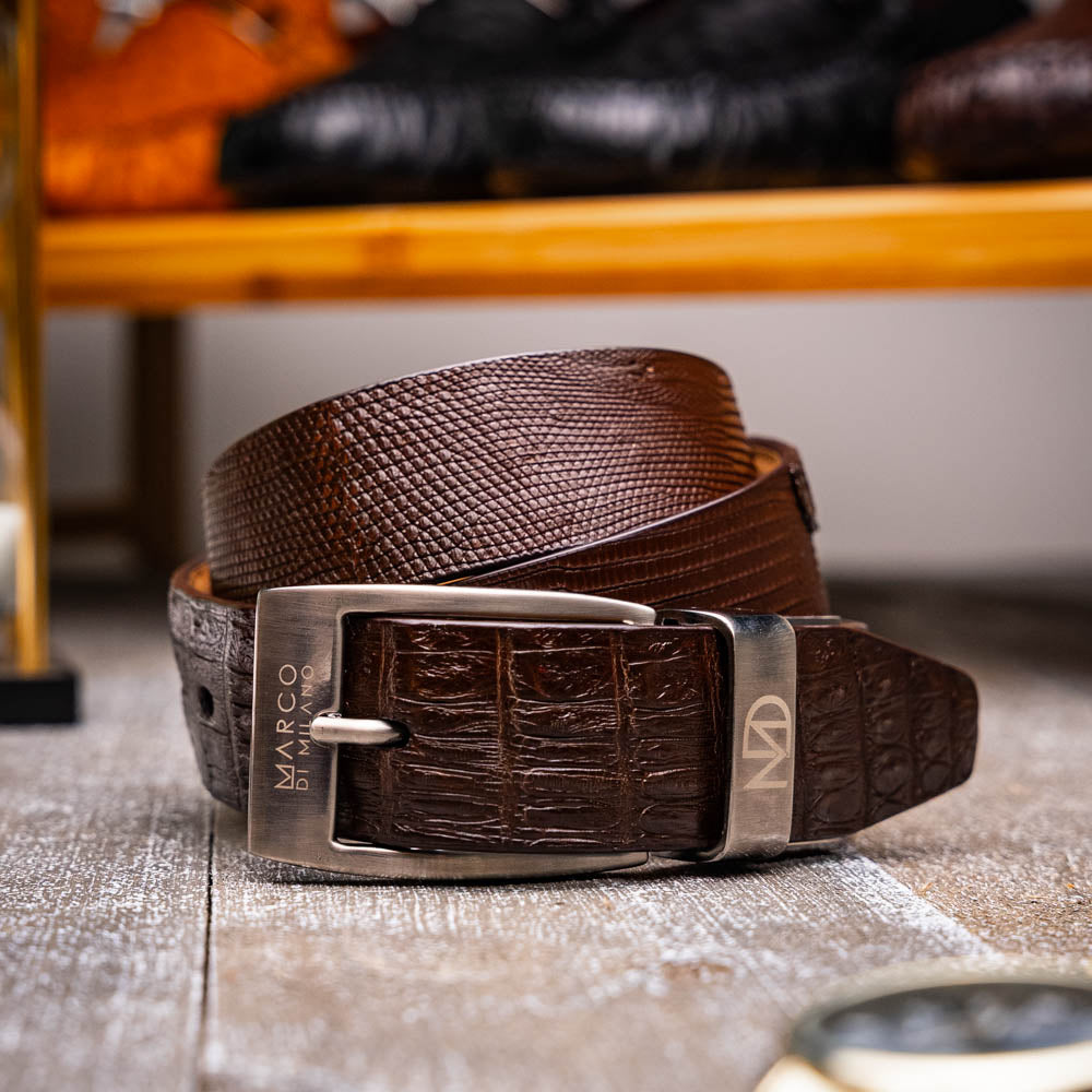 The Giovanni Belt Brown by Marco Di Milano is a dark brown leather belt with a textured pattern, finished with a silver buckle engraved with "Marco" and the "MD" logo, crafted from genuine lizard ring-caiman belly leather.