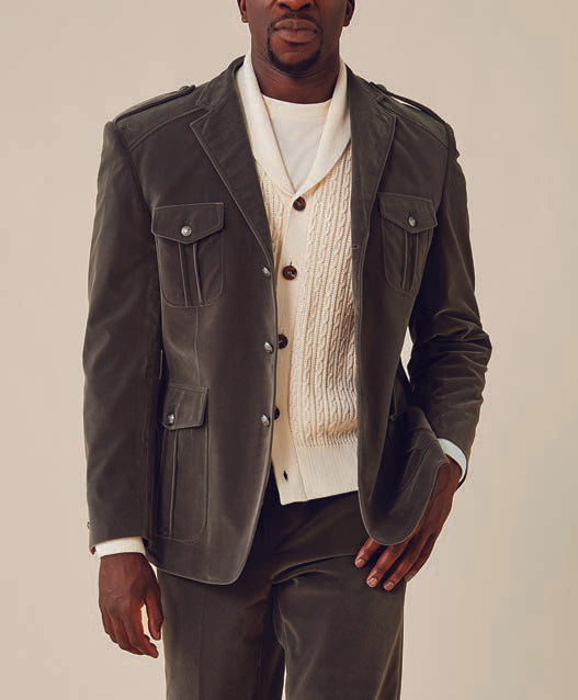 Someone is dressed in an Inserch Velvet Military Blazer in a gray hue, featuring military shank buttons, layered over a cream cable-knit sweater and paired with matching pants. The background is a plain off-white.