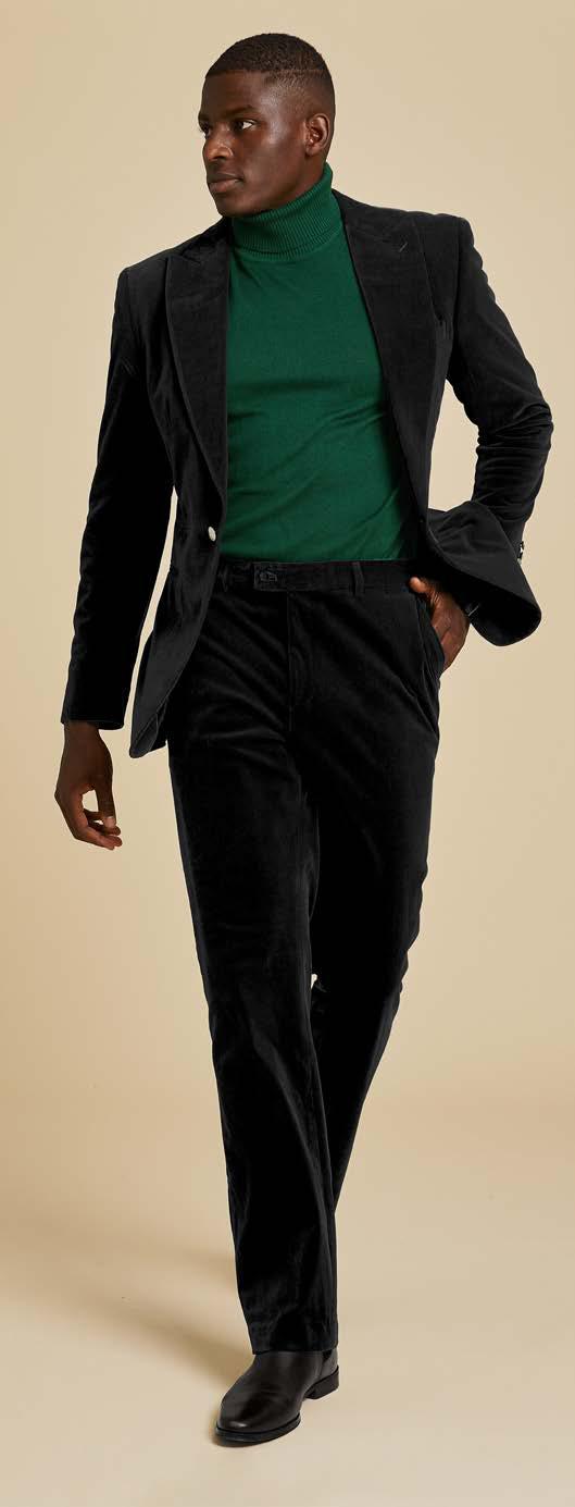 A man stands against a beige background, looking to the side, wearing the Inserch Velvet Blazer BL007-01 Black by Inserch over a green turtleneck with black shoes.
