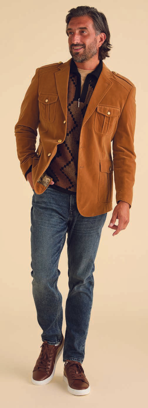 A gentleman wearing the Inserch Velvet Military Blazer BL569-184 in Brown Sugar, accompanied by a patterned sweater, jeans, and brown sneakers, stands and smiles against a beige background.