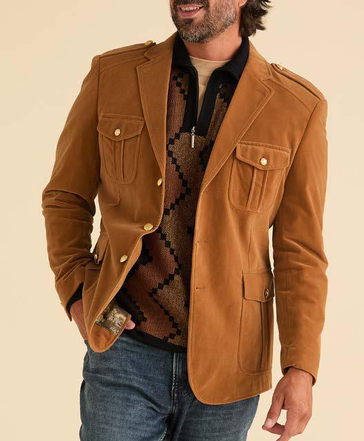A gentleman wearing the Inserch Velvet Military Blazer BL569-184 in Brown Sugar, accompanied by a patterned sweater, jeans, and brown sneakers, stands and smiles against a beige background.