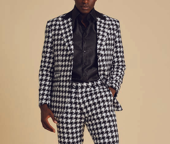 The Inserch Wool Blend Houndstooth Check Peak Lapel Suit in Black/White, from the brand Inserch, creates a sophisticated ensemble with its subtle modern suede trim. Paired with a sleek black shirt, the individual stands confidently with one hand in their pocket against a plain background.