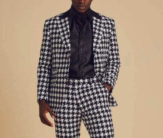 The Inserch Wool Blend Houndstooth Check Peak Lapel Suit in Black/White, from the brand Inserch, creates a sophisticated ensemble with its subtle modern suede trim. Paired with a sleek black shirt, the individual stands confidently with one hand in their pocket against a plain background.