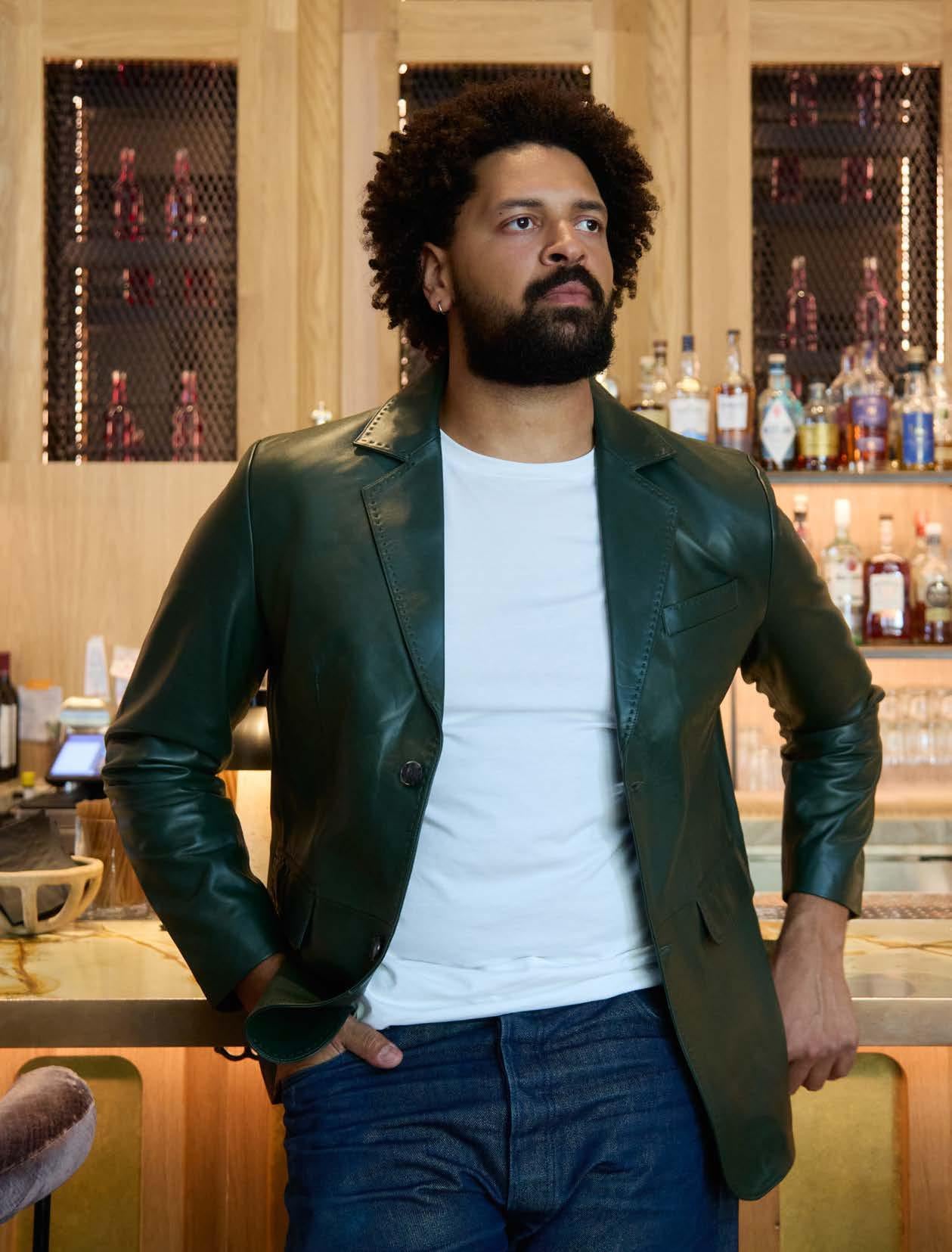 A man with curly hair and a beard wears the Inserch Leather Blazer 600-00052 Pine, featuring pique stitch detail, over a white shirt as he stands by a wooden bar with liquor bottles in the background.