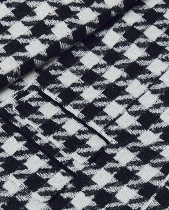 The Inserch Wool Blend Houndstooth Check Peak Lapel Suit in Black/White, from the brand Inserch, creates a sophisticated ensemble with its subtle modern suede trim. Paired with a sleek black shirt, the individual stands confidently with one hand in their pocket against a plain background.