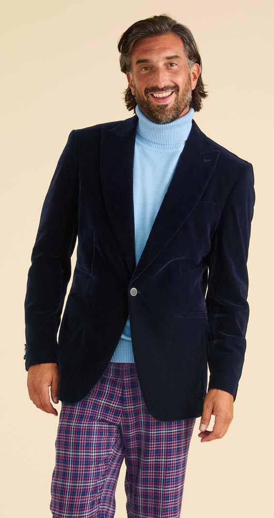 A man smiles against a plain background, wearing the Inserch Velvet Blazer BL007-11 Navy Blue by Inserch with a light blue turtleneck sweater and purple plaid pants, one hand by his side.