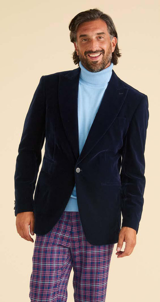 A man smiles against a plain background, wearing the Inserch Velvet Blazer BL007-11 Navy Blue by Inserch with a light blue turtleneck sweater and purple plaid pants, one hand by his side.