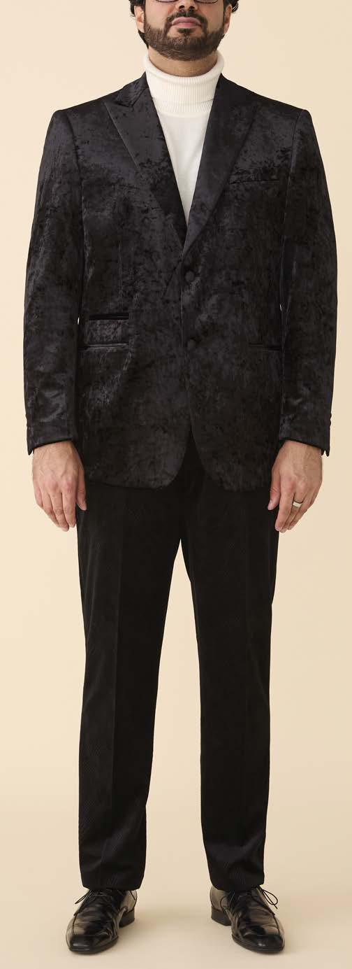 A man wears the Inserch Crushed Velvet Blazer BL008-00001 Black by Inserch over a white turtleneck sweater with black dress shoes, standing against a beige background.