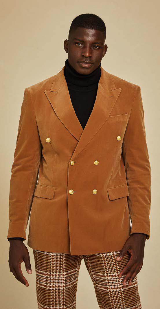 A man wears the Inserch DB Velvet Blazer BL502-184 Brown Sugar with gold buttons, a black turtleneck, and brown plaid pants, standing against a beige background.