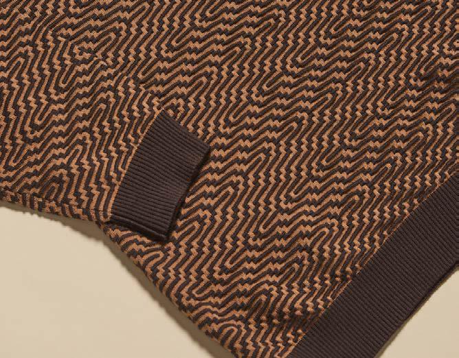 A person wearing the Inserch Intarsia Turtleneck Sweater SW304-00043 Espresso by Inserch, featuring a brown and orange geometric pattern with dark brown accents, paired with dark brown pants, stands against a beige background.