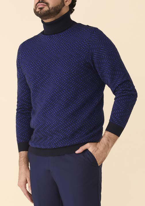 A man wearing the Inserch Intarsia Turtleneck Sweater SW304-00036 Ink by Inserch and navy pants stands against a beige background with one hand in his pocket.