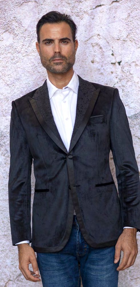 A man wearing a MANZINI MZV-565 Fancy Velvet Sport Coat Black Tailored fit with a patterned lapel, over a white button-down shirt and blue jeans.