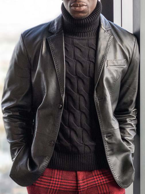 Wearing the Inserch Leather Blazer 600-00001 Black by Inserch—featuring a modern fit and subtle pique stitch detail—a person stands by a window with hands in their pockets. They pair the sleek black lamb leather blazer with a black cable-knit turtleneck sweater and striking red plaid pants.