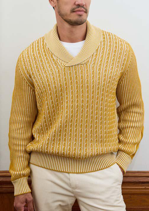 A man wearing the Inserch Shawl Collar Cable Sweater SW005-00038 Gold—a yellow and white patterned cotton acrylic blend sweater by Inserch featuring a shawl collar—paired with cream-colored pants, stands against a plain wall with one hand in his pocket.
