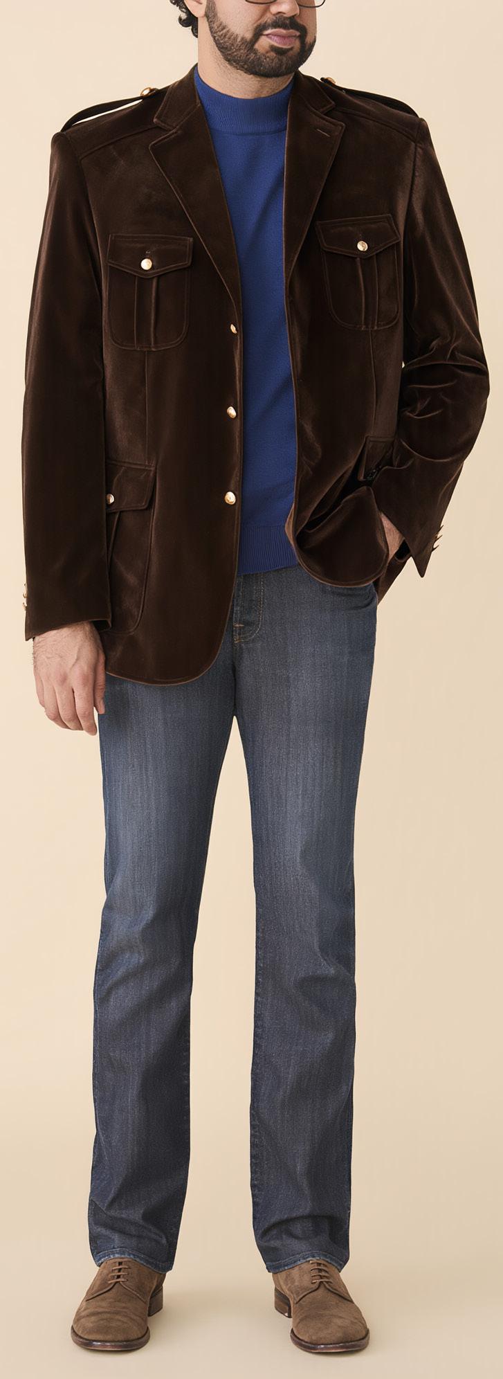 A man wearing the Inserch Velvet Military Blazer BL569-00024 Chocolate by Inserch, paired with a blue sweater, blue jeans, and brown shoes, stands against a beige background.