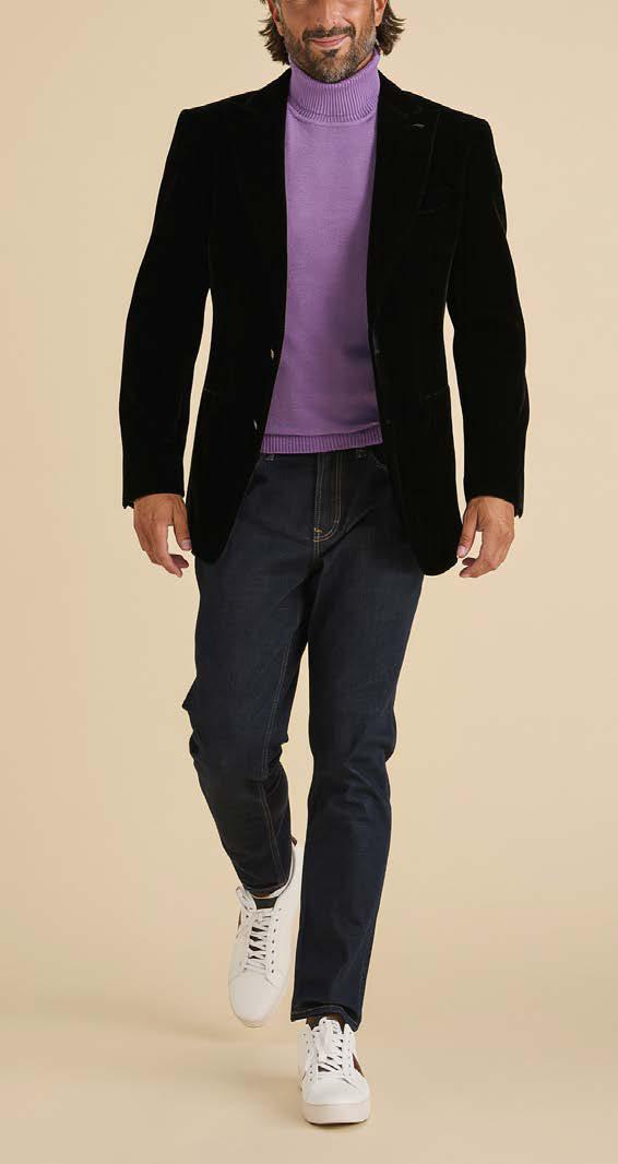 A man wearing the Inserch Velvet Blazer BL007-00001 Black by Inserch, featuring a peak lapel and shank buttons, paired with a purple turtleneck sweater, dark jeans, and white sneakers, walks forward against a beige background.