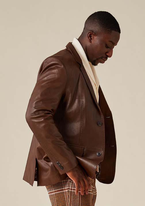 A man wearing the Inserch Leather Blazer 600-00104 in Cognac by Inserch—crafted from brown lamb leather with subtle pique stitch detailing—paired with a cream shirt and plaid pants, stands against a plain beige background, looking down with one hand by his side; the blazer’s paisley print lining offers a refined finishing touch.