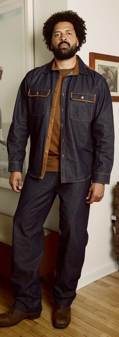 A man stands against a beige background wearing the Inserch Denim Shirt w/ Suede Trim 2pc Set in indigo by Inserch, paired with an orange sweater and dark brown shoes.
