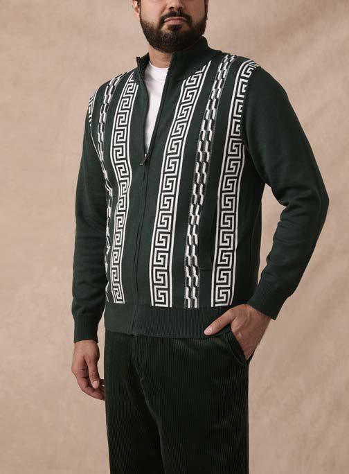A man wearing the Inserch Greek Cable Full Zip Sweater SW617-00021 in Hunter Green by Inserch, featuring white Greek key patterns, styled over a white shirt and green corduroy pants, stands against a beige background.