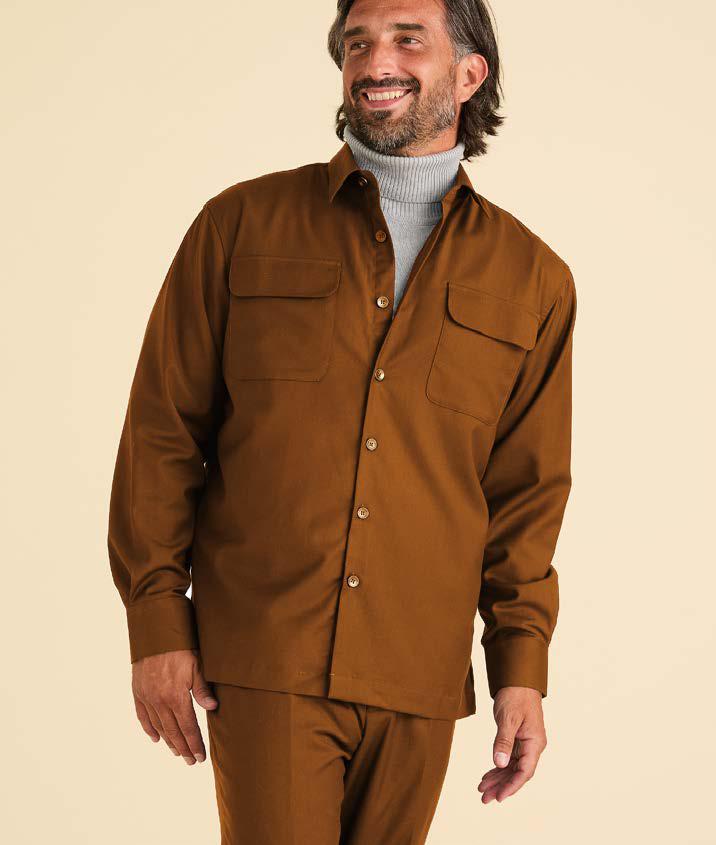 A man wearing the Inserch Two-Pocket Shirt SE004-00027 Aztec by Inserch—brown, regular fit, long sleeve with two pockets—over a gray turtleneck and matching brown pants stands against a plain light background, smiling and looking to the side.