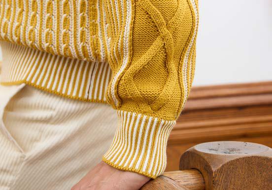 A man wearing the Inserch Shawl Collar Cable Sweater SW005-00038 Gold—a yellow and white patterned cotton acrylic blend sweater by Inserch featuring a shawl collar—paired with cream-colored pants, stands against a plain wall with one hand in his pocket.