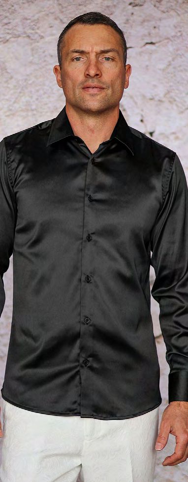 A man stands before a light textured background wearing the Manzini INR-101 Slim Fit Satin Spandex Black Shirt, paired with white pants.