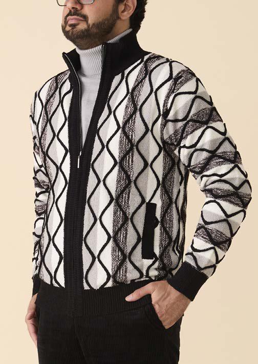 A man wears the Inserch Bouclé Yarn Graphic Full Zip Sweater (SW614-00001) in black—featuring a bold graphic knit pattern and a metal zipper closure from Inserch—layered over a light turtleneck and paired with dark pants, standing with one hand in his pocket.