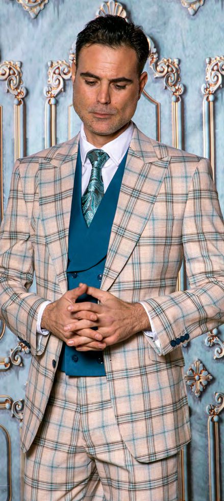 A man wearing the Insomnia PARIS-5 3PC Compose Tailored Fit Teal Suit by Insomnia stands with hands clasped, looking down against an ornate decorative background.